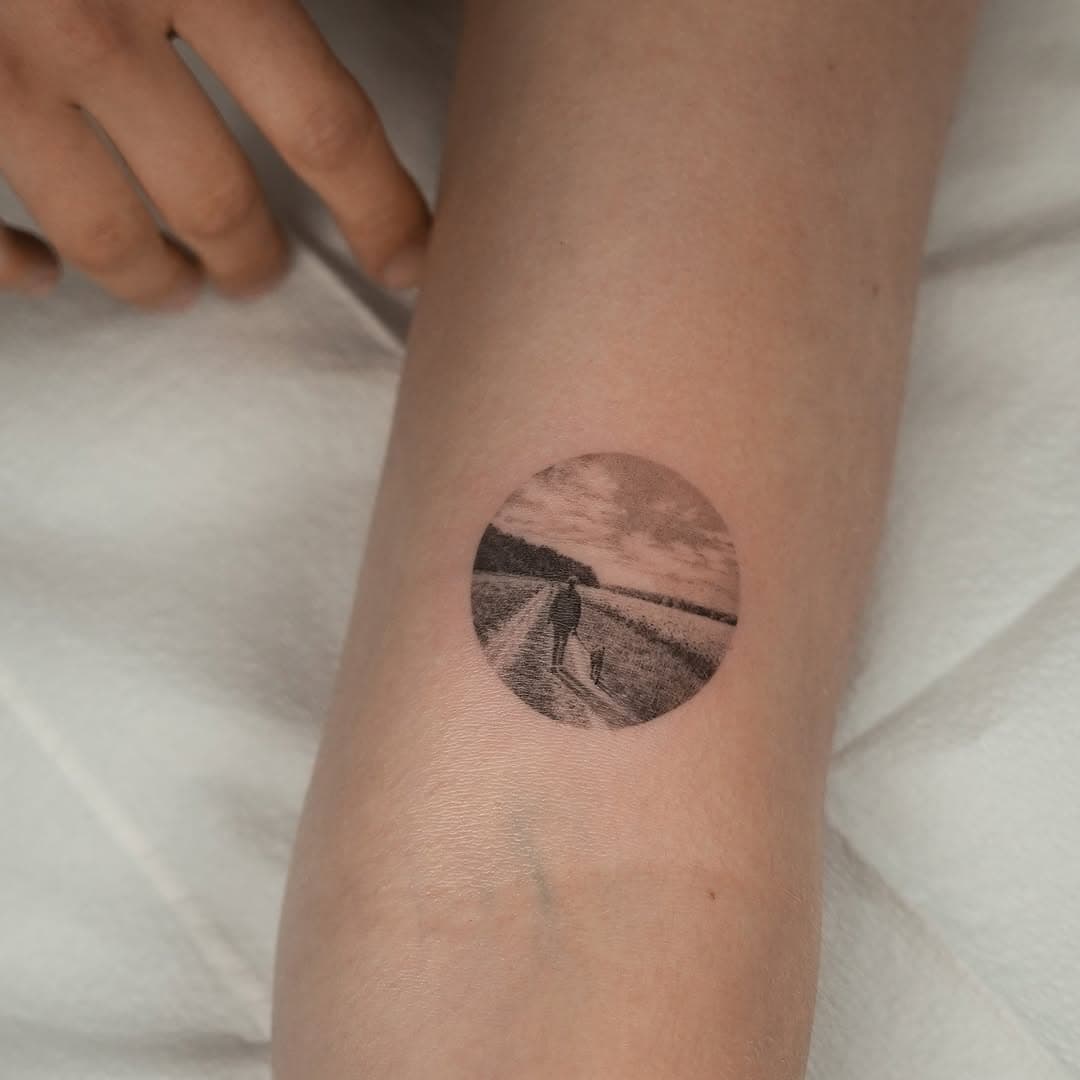 Small circular tattoo on wrist depicting a solitary figure walking on a rural path through fields in fine-line black ink style