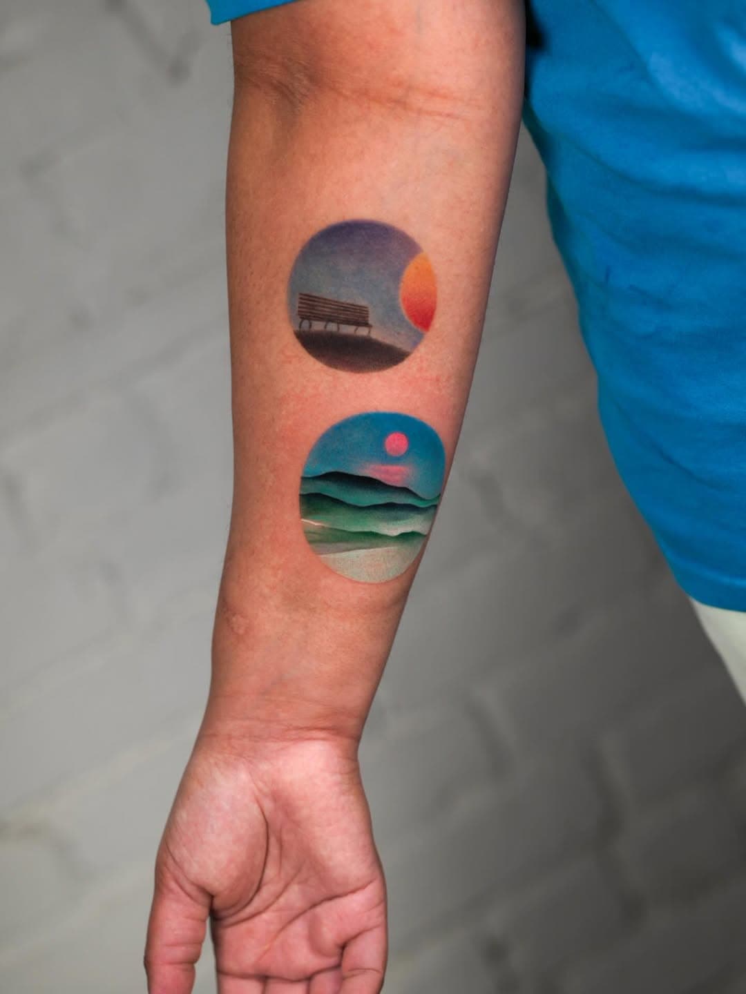 Two circular landscape tattoos on forearm featuring minimalist scenes with sun/moon, ocean waves, and layered mountain horizons in blue and orange ton