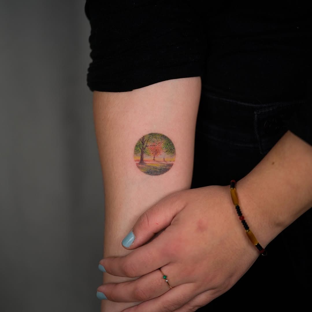 Small circular tattoo on arm showing colorful landscape scene with trees in impressionist style, featuring pink and green tones