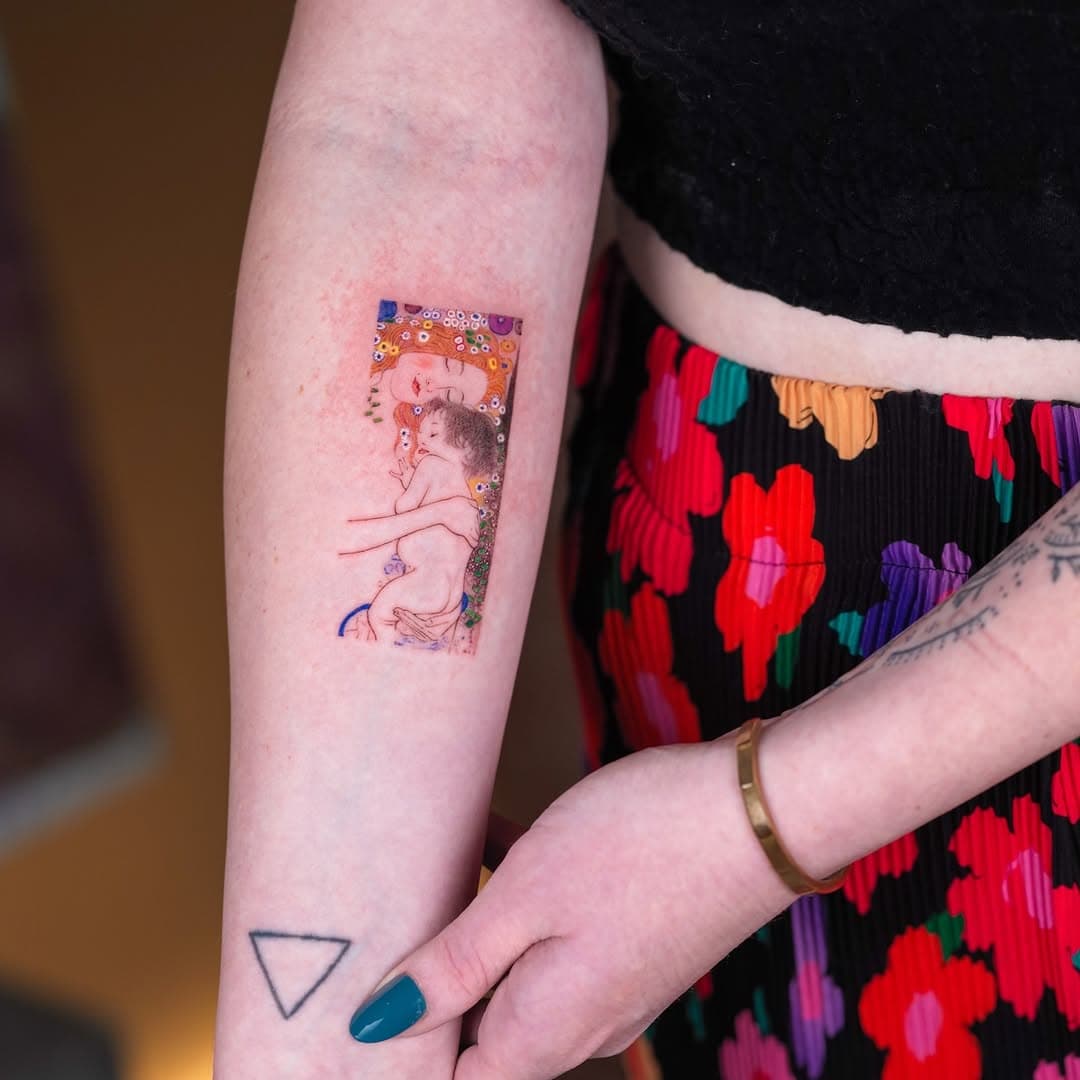 Colorful illustrative tattoo of a classical nude figure surrounded by ornate decorative patterns and jewel-toned embellishments on upper arm