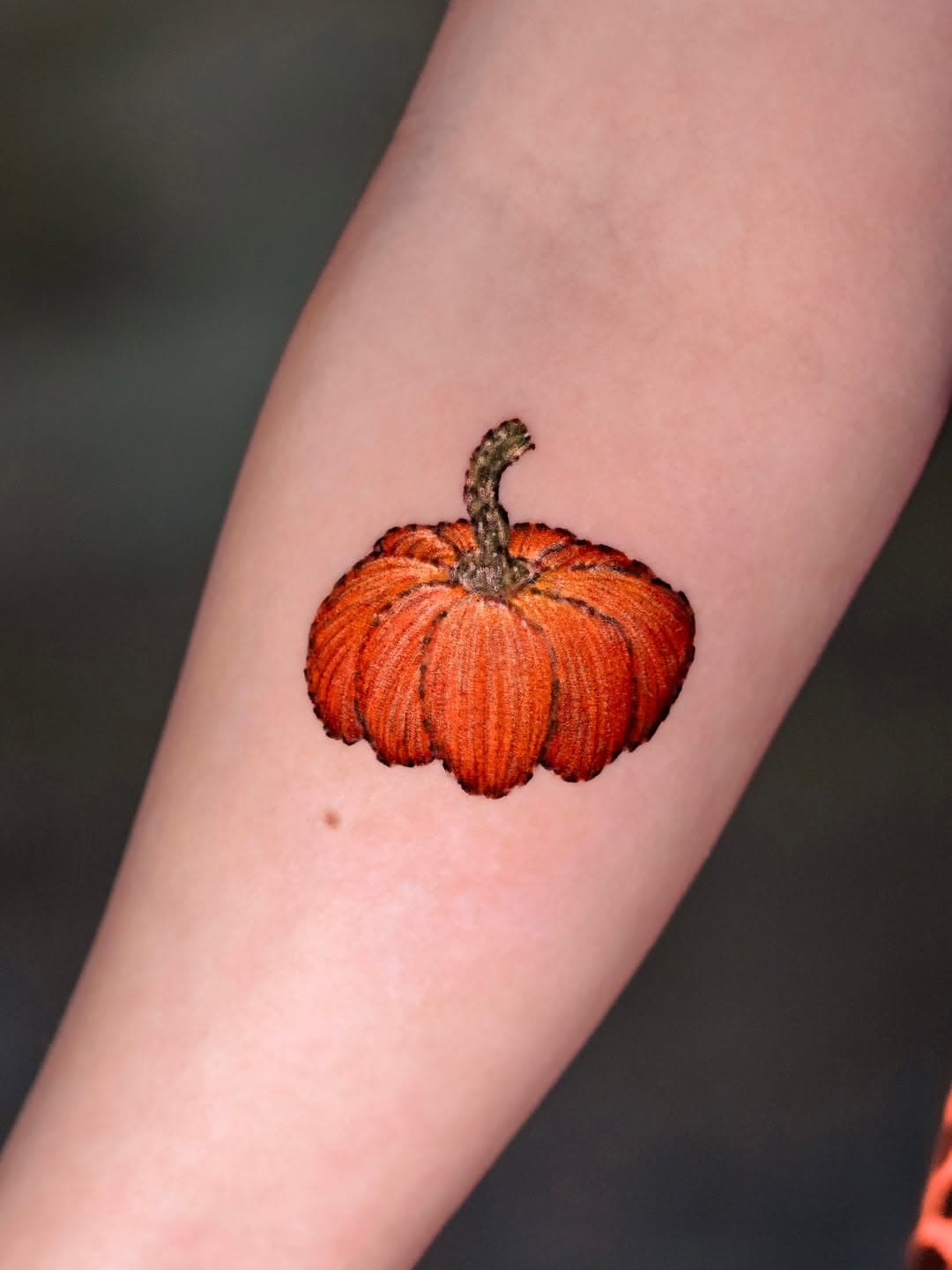 Small illustrative pumpkin tattoo with orange and red shading on forearm, featuring curved stem and textured ridges in surrealist style