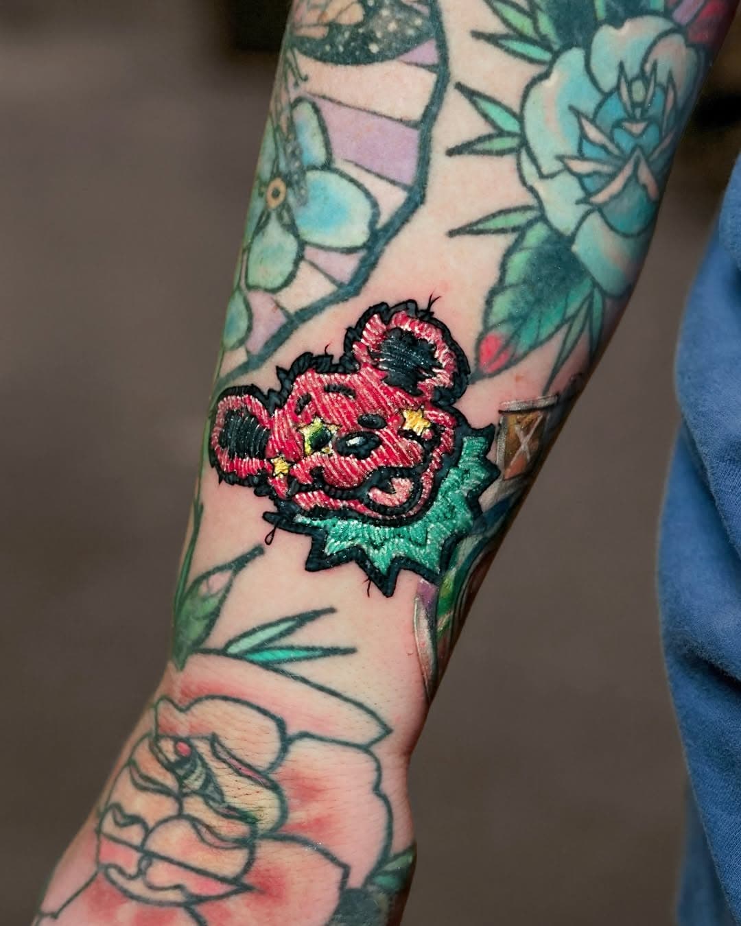 Colorful embroidered patch-style Minnie Mouse tattoo on forearm surrounded by floral designs in illustrative surrealism style