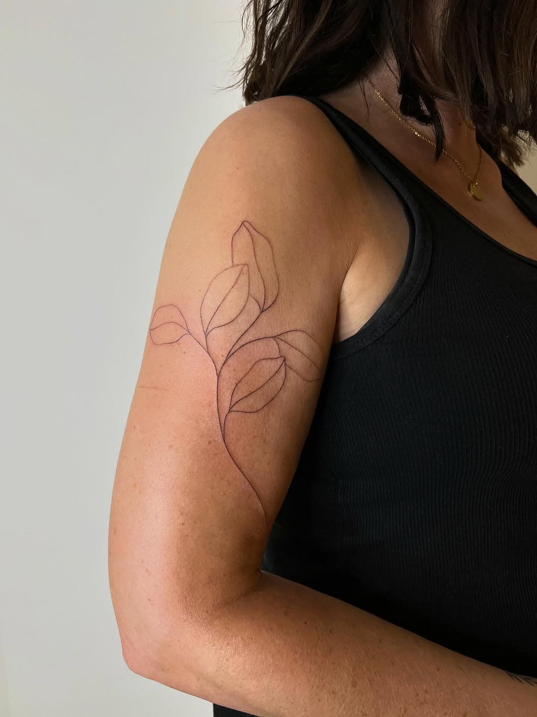 Fine-line minimalist tattoo of botanical leaves with delicate outlines on upper arm by Lara Maju