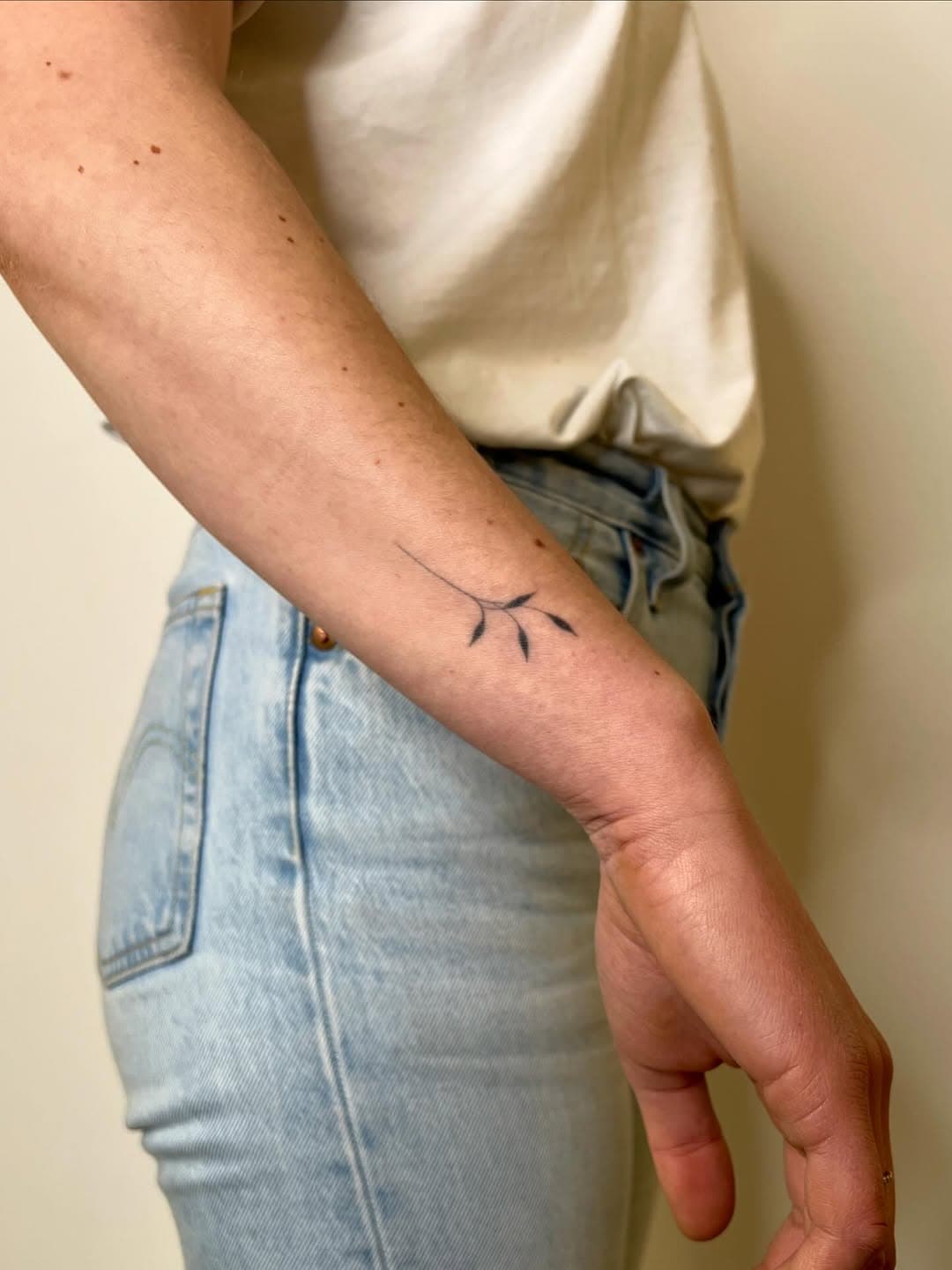 Minimalist fine-line tattoo of a simple branch with leaves on inner forearm