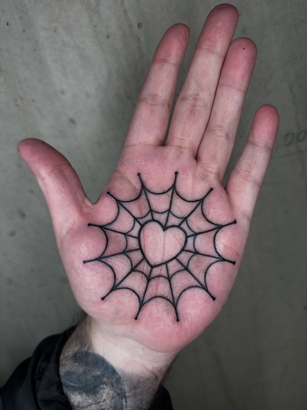 A black fine-line spider web tattoo centered on an open palm, drawn in minimalist illustrative style with radiating geometric lines