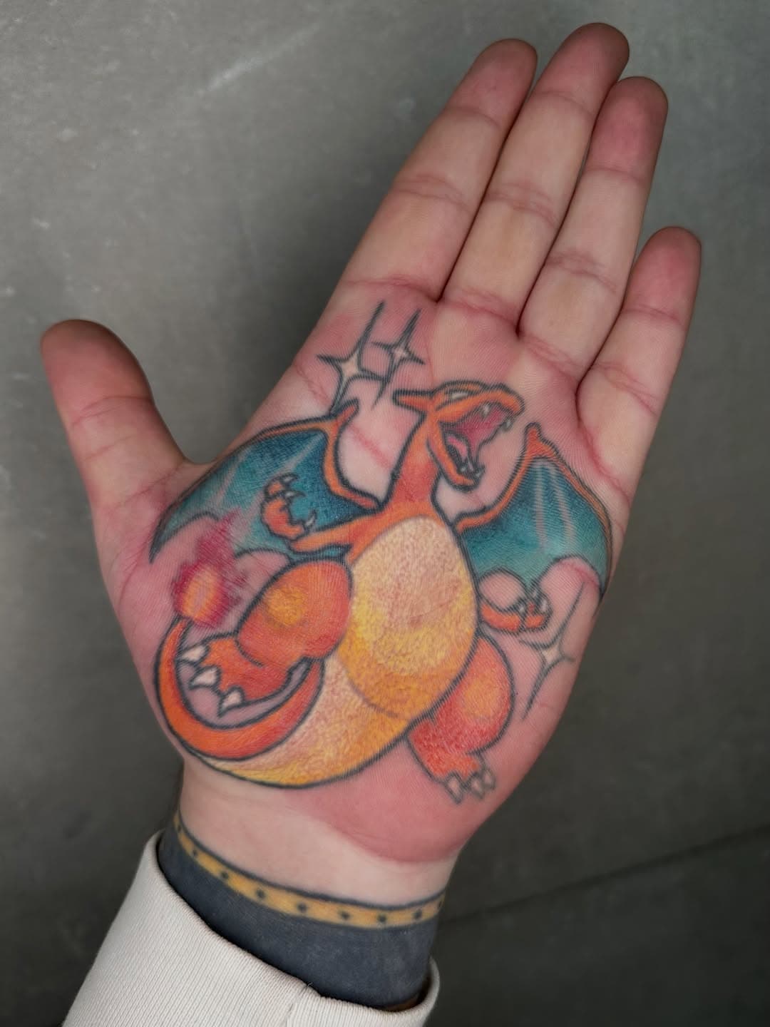 Colorful illustrative palm tattoo of Charizard Pokemon with orange body, blue-green wings, and flame tail in fine-line style