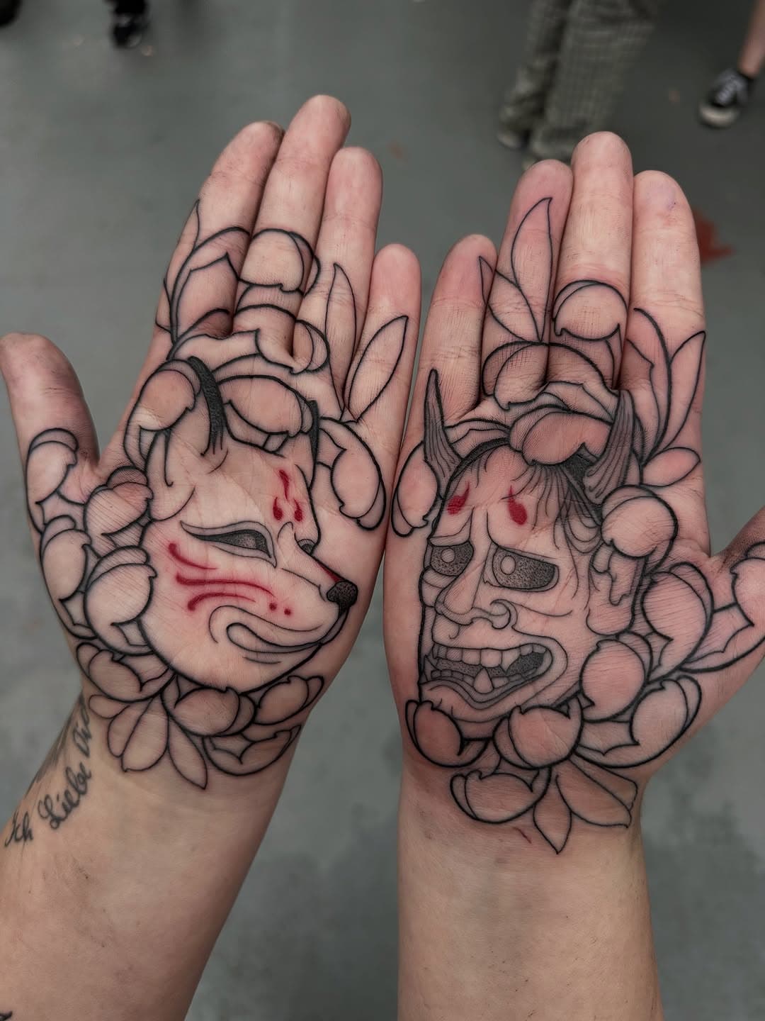 Two palms with matching Japanese-inspired tattoos featuring Hannya mask and Kitsune fox mask surrounded by flowing peony flowers in black linework