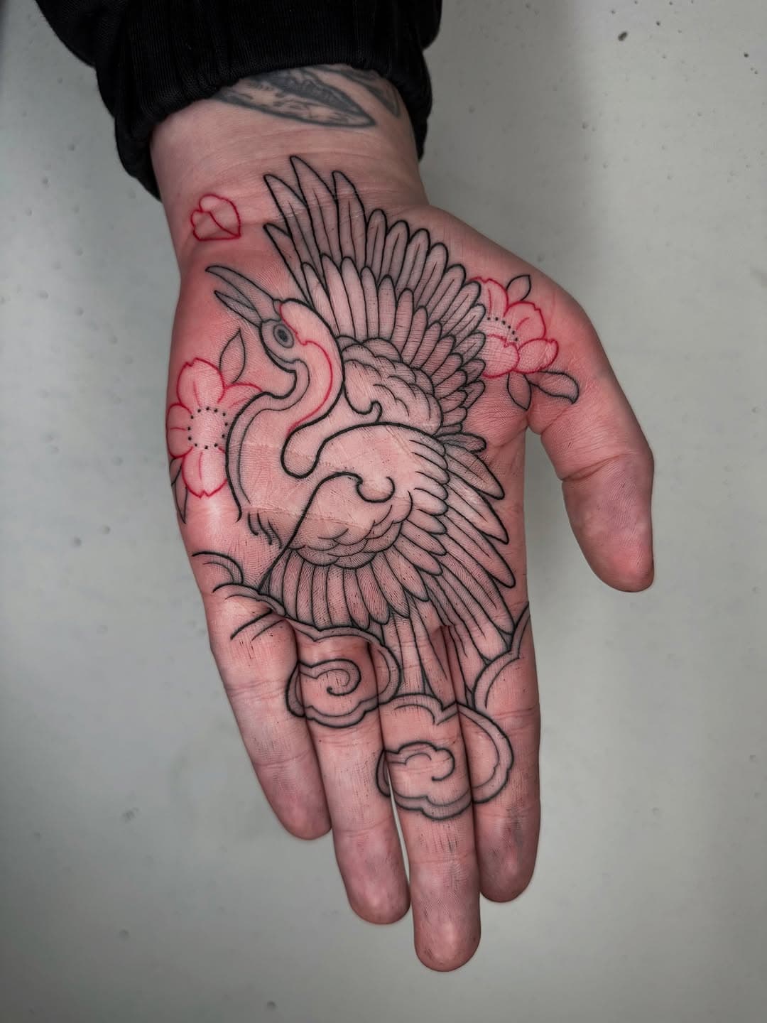 Black line tattoo of a crane bird with spread wings and red floral accents covering an open palm in illustrative style