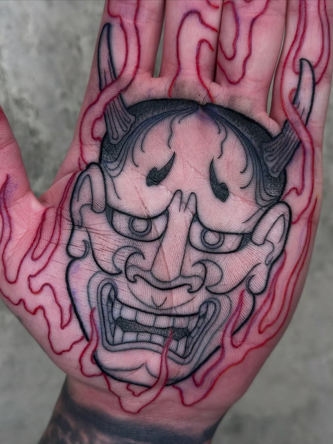 Hand tattoo of a Japanese oni mask in black linework surrounded by red flame-like design extending to fingers