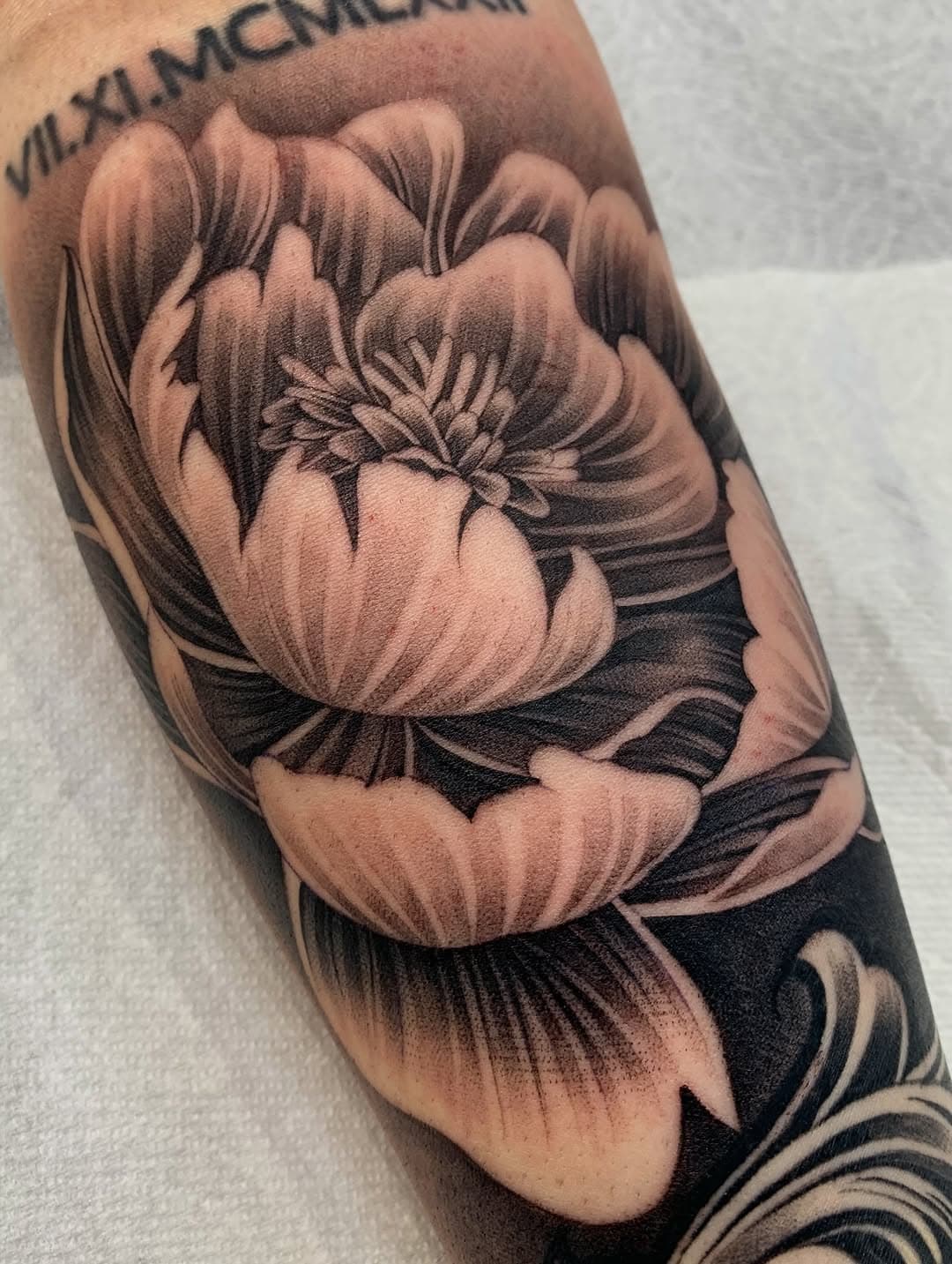 Black and grey realistic peony flower tattoo with layered petals and dark shading on forearm by Adrian Castro