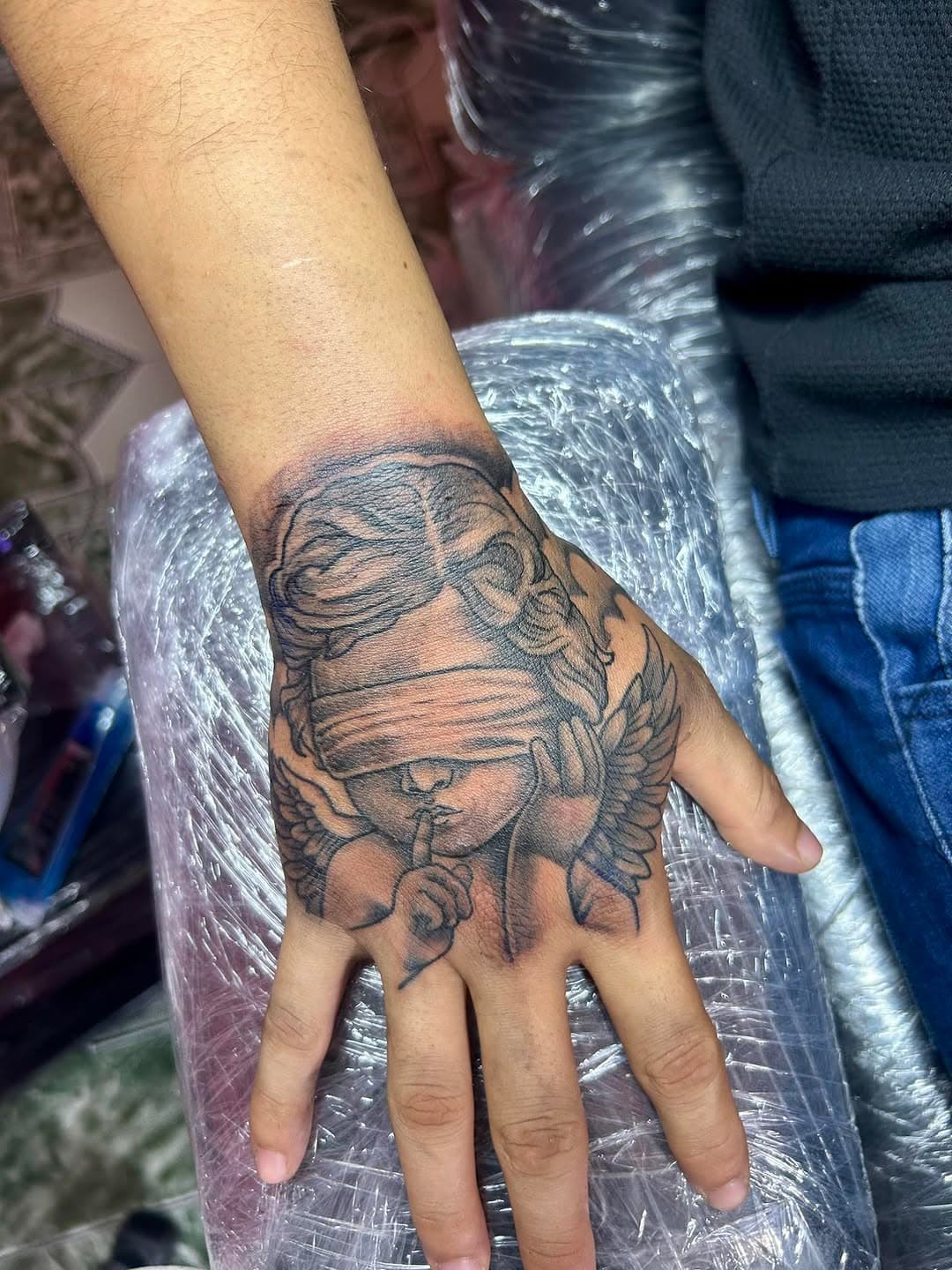 Hand tattoo featuring a blindfolded angel with wings in a shushing gesture, done in fine-line black and grey style