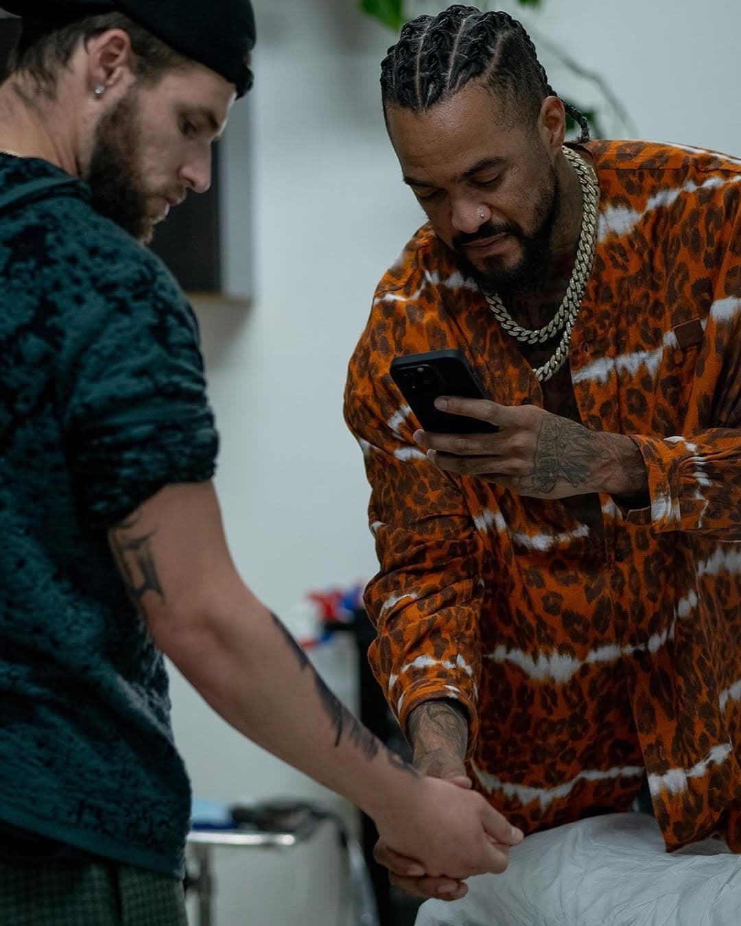 Artist tattooing client's forearm in black and grey while client in orange leopard print outfit checks phone