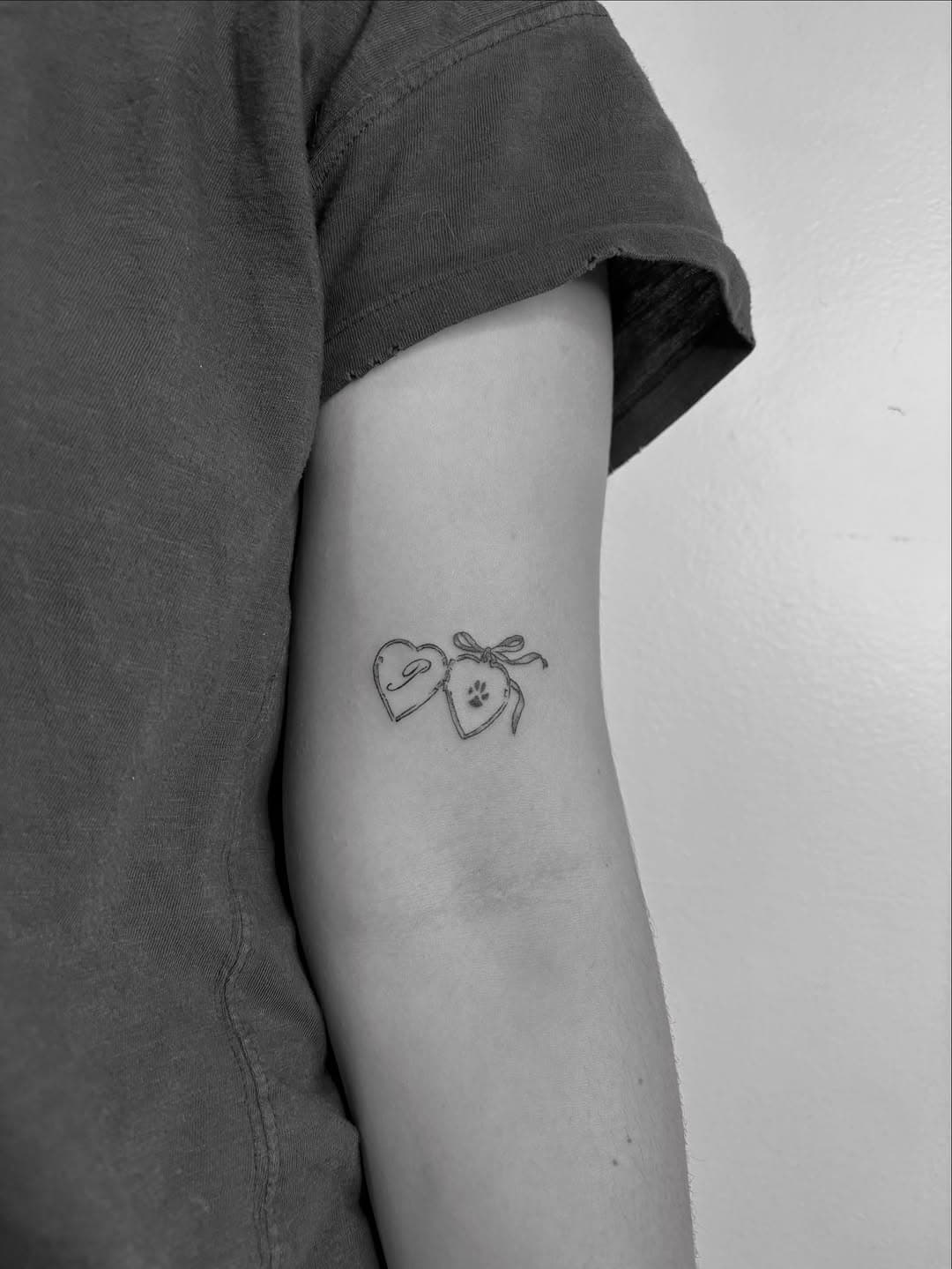 Fine-line tattoo of two heart lockets connected by a bow, one containing a dog silhouette and one with a paw print, on upper arm