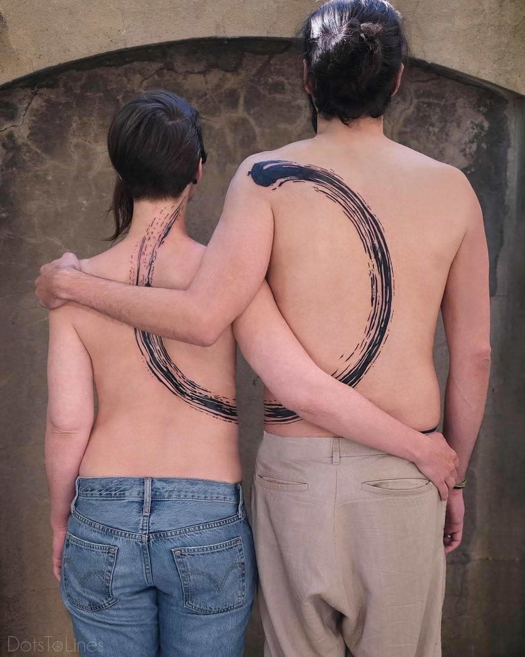 Two people showing matching brushstroke circular tattoos on their backs that connect when standing together