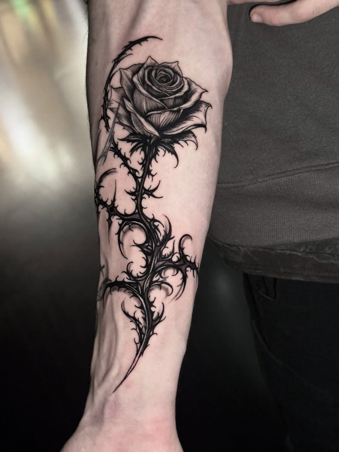 Black and grey illustrative rose tattoo with ornate thorny stem on forearm in decorative style